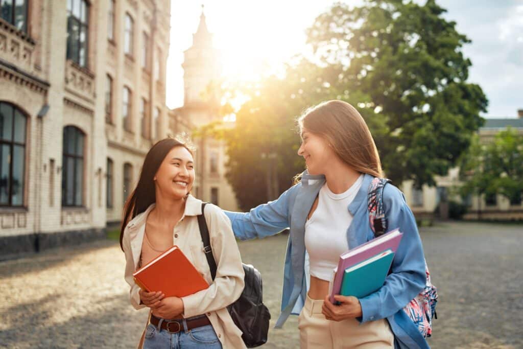 Two joyful female students walking together on campus holdi 1024x683